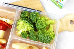 Broccoli florets in a clear lunch container on a wooden surface with bread slices nearby and a tomato piece visible in the corner.