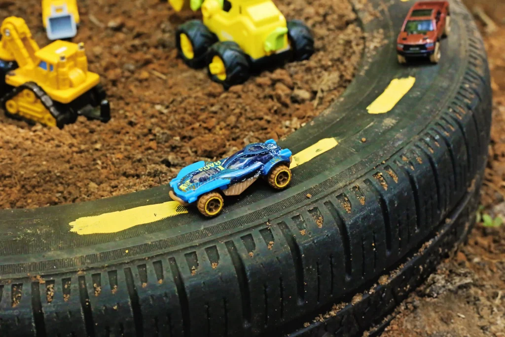 Blue toy race car speeds along a curved black track on dirt, with a yellow construction toy and a red toy truck in the background for this car race track activity