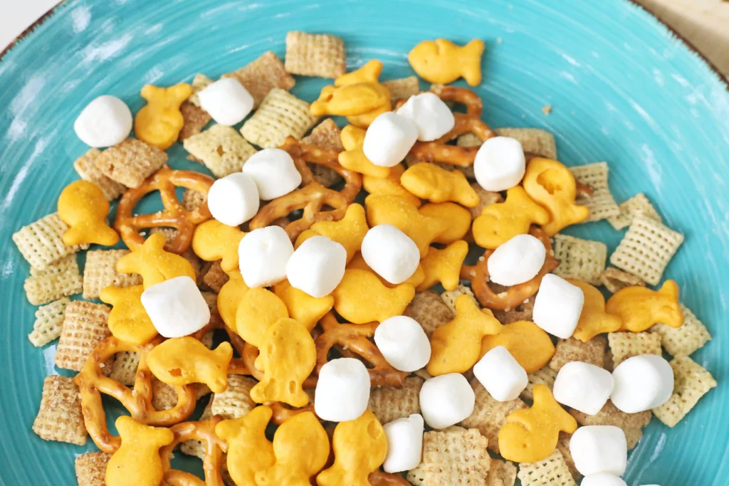 Snack mix with cereal squares, pretzels, goldfish crackers, and white marshmallows on a turquoise plate.