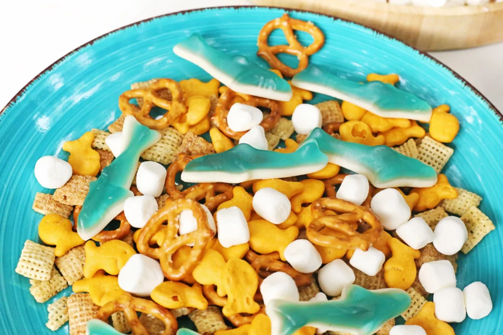 Turquoise plate of snack mix with gummy sharks, pretzels, mini marshmallows, and cereal pieces.