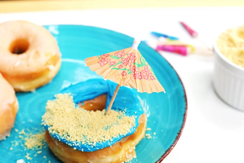 Donut with bright blue glaze and a small decorative paper umbrella on a turquoise plate, crumbs around the glaze.