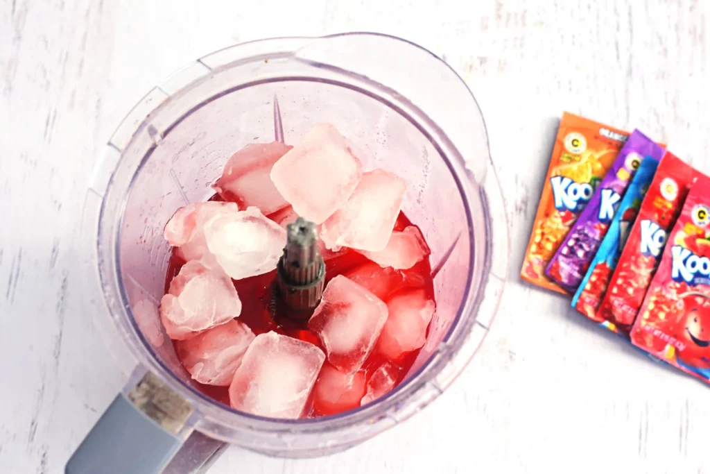 Kool-Aid Slushie - Cultivating Brilliant Minds Blender jar filled with ice chunks and red kool aid and water, ready to blend, with colorful kool aid packets lying to the right.