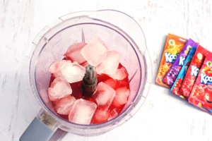Blender jar filled with ice chunks and red kool aid and water, ready to blend, with colorful kool aid packets lying to the right.