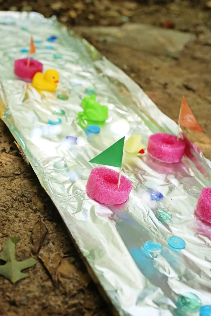 Aluminum Foil River - Cultivating Brilliant Minds aluminum foil river with pink pool noodle slices, a yellow rubber duck, and colorful paper flags on the ground
