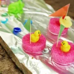 Pink pool noodle slices with yellow rubber duck toppers on skewers arranged on aluminum foil near a sandy surface