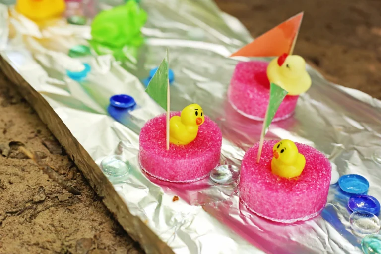 Pink pool noodle slices with yellow rubber duck toppers on skewers arranged on aluminum foil near a sandy surface