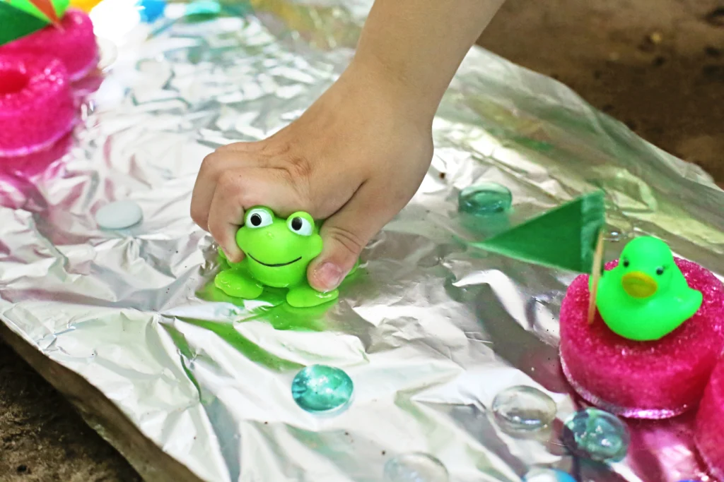 Aluminum Foil River - Cultivating Brilliant Minds Child's hand presses a green frog on a shiny foil surface.