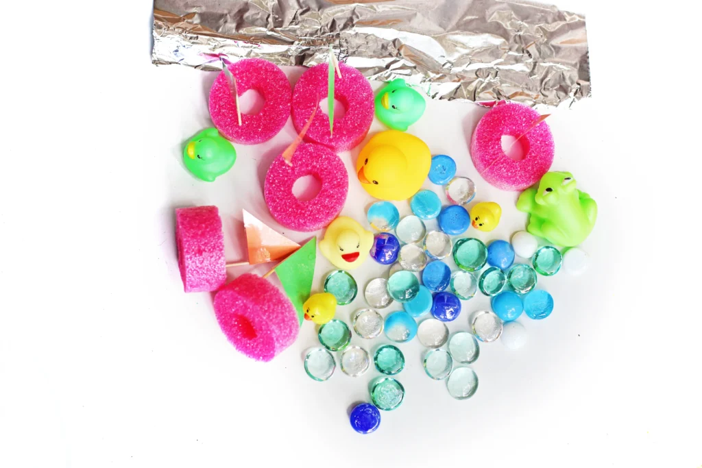 Aluminum Foil River - Cultivating Brilliant Minds supplies for this water play activity for kids - pink pool noodle slices, rubber ducks, bright marbles, and foil on a white background.