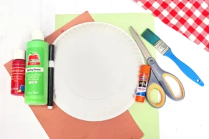 supplies for this apple core craft: red and green acrylic paints, a black marker, scissors, glue, a paper plate, ruler and brushes on colorful backing papers.