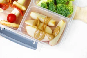 Sectioned lunch container with apple slices spread with peanut butter, broccoli florets, and a cherry tomato for a healthy snack.