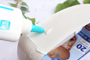 Close-up of a white glue bottle squeezing glue onto a white surface, with a blue nozzle visible.