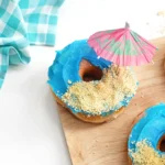 poolside snacks that consist of glazed donuts with blue icing, crushed oreos for sand and a mini umbrella