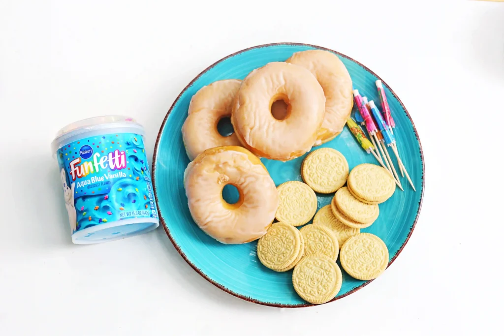 ingredients for the beach donuts - Turquoise plate filled with glazed donuts and round sandwich cookies, with a Funfetti container nearby for a festive snack display.
