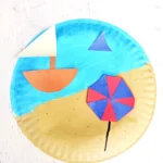 a Beach Paper Plate Craft that is a paper plate painted half blue half tan with construction paper umbrella and sail boat