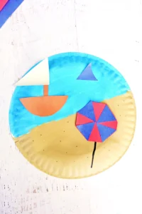 a Beach Paper Plate Craft that is a paper plate painted half blue half tan with construction paper umbrella and sail boat