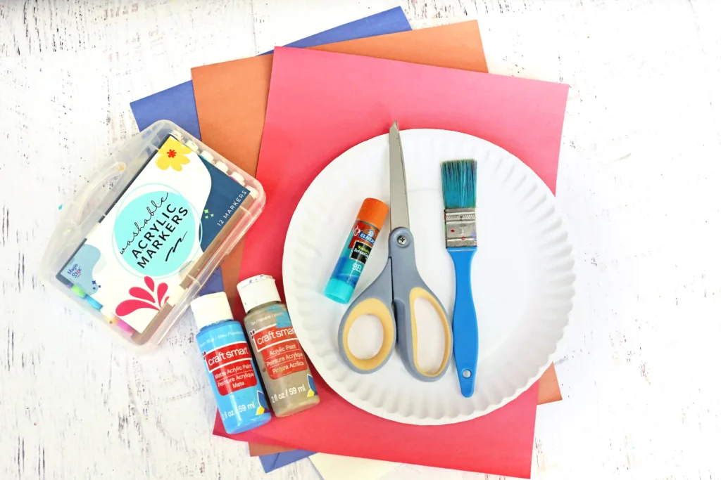 supplies for the beach paper plate craft- Flat lay of art supplies on a white wood table: colored construction paper, bottles of acrylic paint, scissors, a paintbrush, and glue tubes.