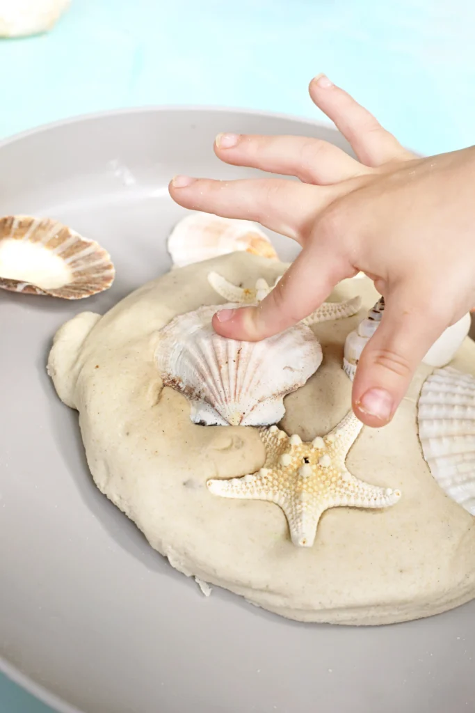 Hand arranging or pressing seashells and starfish on a sandy surface inside a bowl; light blue background.