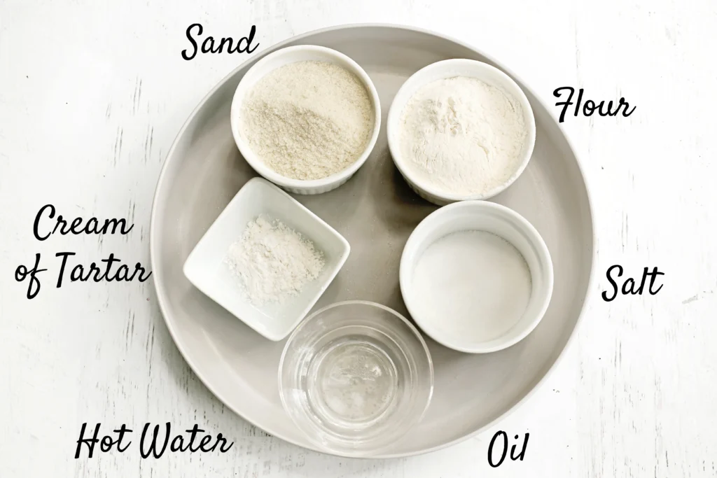 Top-down view of a metal tray with small bowls holding ingredients labeled Sand, Flour, Salt, Hot Water, and Oil on a white wooden surface.