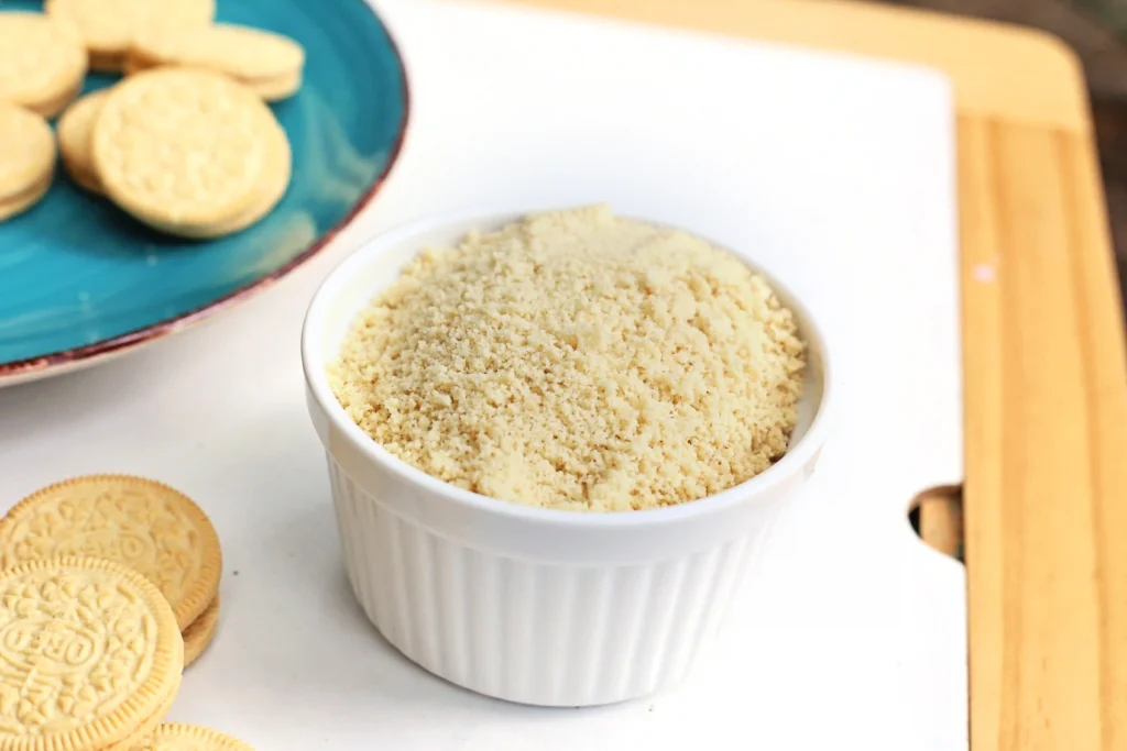 crushed vanilla oreos in a white ramekin with cookies on a blue plate nearby on a white surface.