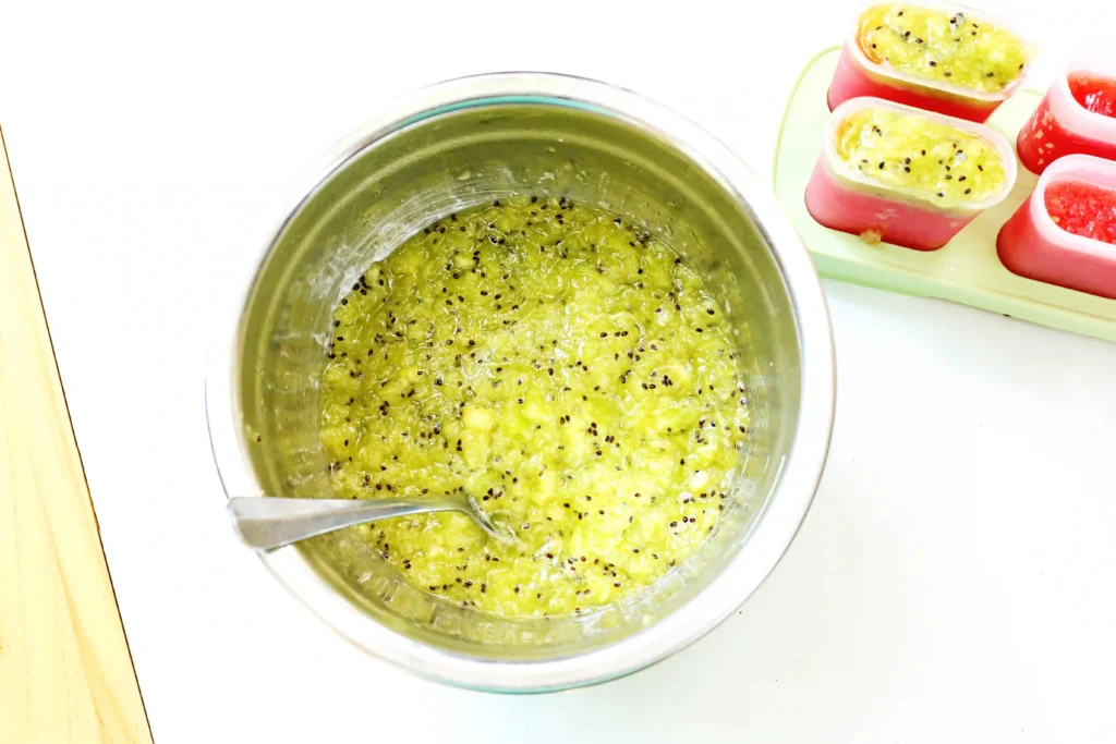 Watermelon Kiwi Popsicles - Cultivating Brilliant Minds Pot with mashed green fruit (kiwi) and black seeds shown with a spoon nearby and small kiwi-topped popsicles on the right.