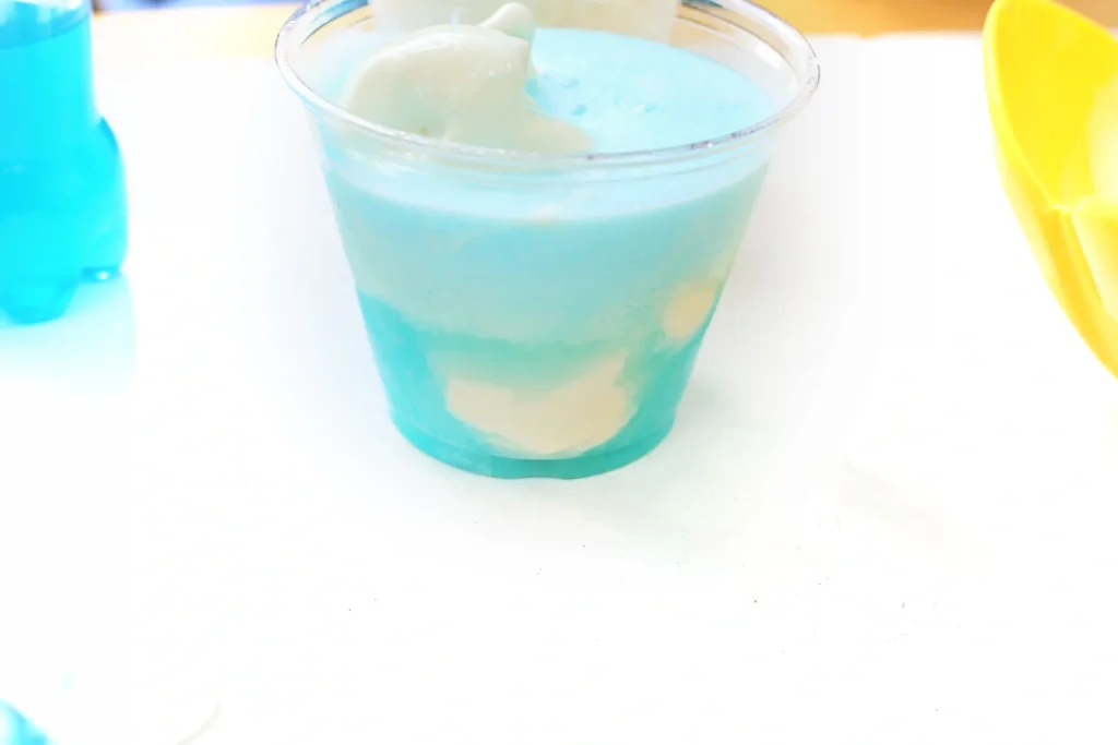 Blue drink and ice cream in a clear cup with ice and lime slices on a white surface.