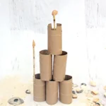 Towering sculpture built from stacked cardboard toilet-paper tubes, with a small shell perched on a vertical stick at the top, on a light surface with seashells nearby