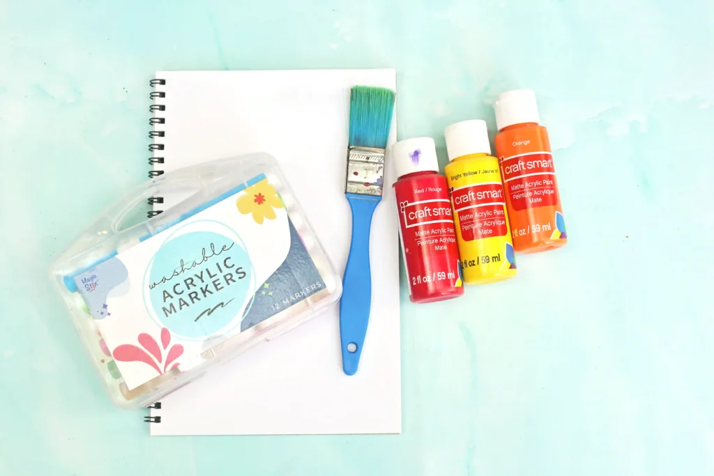 supplies for this alphabet craft- Flat lay of art supplies: notebook, blue paintbrush, acrylic paint bottles (red, yellow, orange) labeled Craft Smart, and a case of washable acrylic markers on a pale blue background
