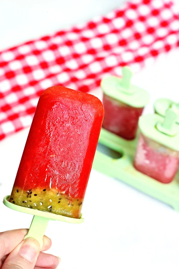 Watermelon Kiwi Popsicles - Cultivating Brilliant Minds Close-up of a bright red popsicle on a light green stick, with kiwi pieces and another green-popped treat in the background on a red checkered backdrop.