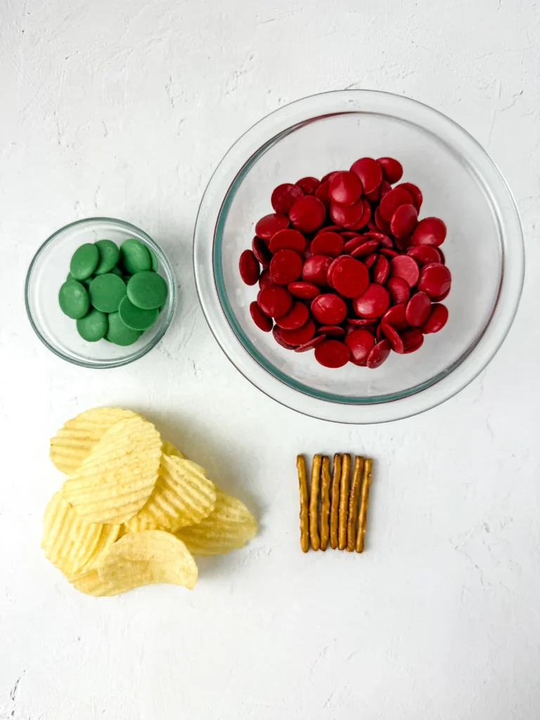 Chocolate Covered Apple Chips - Cultivating Brilliant Minds ingredients for these chocolate covered apple chips - a large glass bowl filled with red candy-coated chocolates, a smaller bowl with green candies, plus potato chips and pretzel sticks nearby.