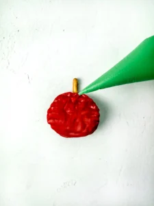 Close-up of a red chocolate covered chip with a small pretzel stem on a white textured surface. A piping bag filled with green melted chocolate is being held over it for this back to school dessert board