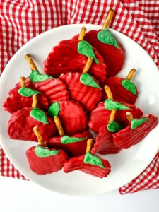Chocolate Covered Apple Chips that are chips covered with red melting chocolate with a pretzel rod for a stem and a green melting chocolate leaf on a white plate for a back to school dessert board