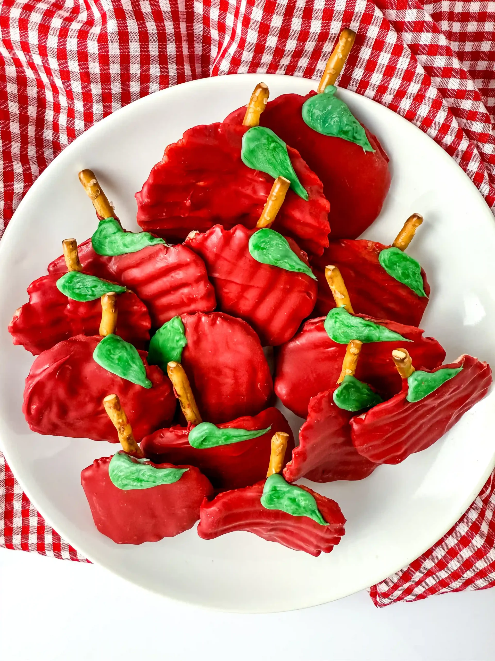 Chocolate Covered Apple Chips that are chips covered with red melting chocolate with a pretzel rod for a stem and a green melting chocolate leaf on a white plate for a back to school dessert board