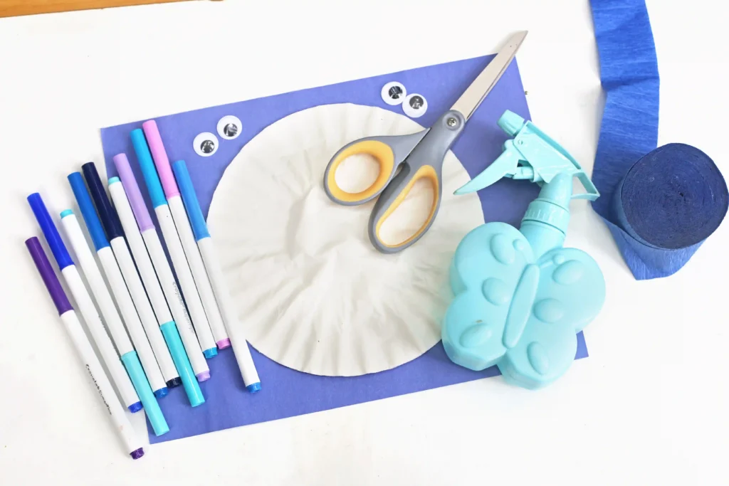 Coffee Filter Jellyfish Craft - Cultivating Brilliant Minds Craft supplies laid out on a white surface: colorful markers, scissors, googly eyes, glue spray, blue paper, and a blue dinosaur-shaped playdough tool wearing a purple-themed project backdrop.