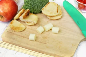 Cutting board scene with a red apple, broccoli, stacked flatbreads, cheese cubes, and a green-handled knife ready for prepping.