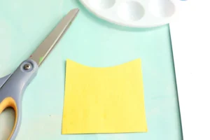 Yellow construction paper cut into the shape of a crayon box on a light blue workspace with scissors and a craft knife nearby.