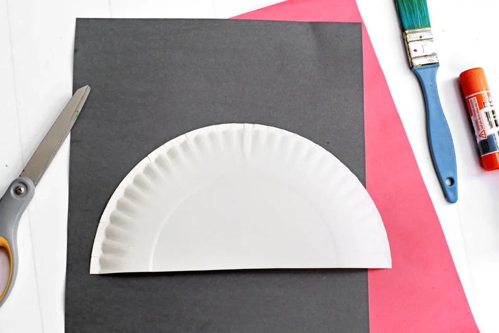 Craft workspace showing dark gray cardstock with a white plate cut in half, a pink paper strip, and art supplies (blue paintbrush, orange glue stick, scissors) laid out on a white surface.