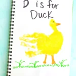 Notebook page with handwriting 'D is for Duck' beside a yellow duck illustration on green grass and a turquoise background.
