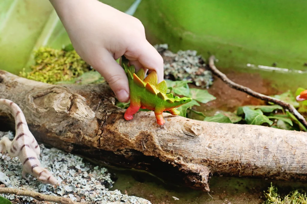 Dinosaur Swamp Water Table - Cultivating Brilliant Minds Child’s hand picking up a green and orange toy dinosaur from a log in a forest setting in a water table activity