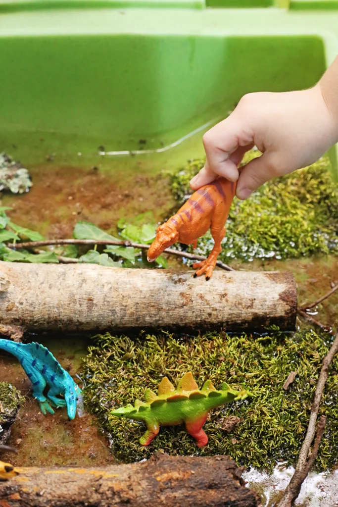 Dinosaur Swamp Water Table - Cultivating Brilliant Minds Close-up of a child's hand gripping an orange toy dinosaur above a mossy surface in a water table for a water play activity