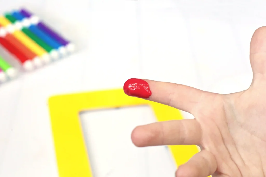Close-up of a red-painted finger near a bright yellow object, with colored pencils blurred in the background.