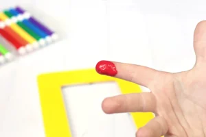 Close-up of a red-painted finger near a bright yellow object, with colored pencils blurred in the background.