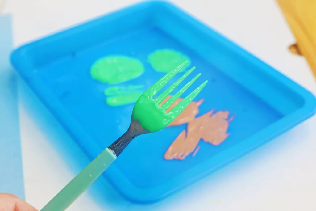 Green plastic fork dipped in bright green paint over a blue paint tray with an orange design in the background.