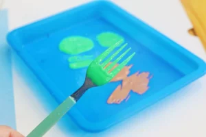 Green plastic fork dipped in bright green paint over a blue paint tray with an orange design in the background.