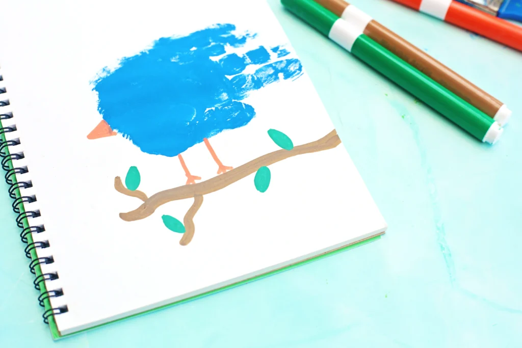 Open notebook page with a blue bird and a simple brown branch drawing, alongside green and brown markers on a pastel blue background for this alphabet craft for kids.