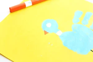 Close-up of a yellow paper with blue paint handprint decorated to look like a pigeon and an orange marker along the top edge for this Mo Willems pigeon craft