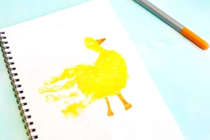 Spiral notebook on a pale blue surface with a yellow handprint paint smear made to look like a duck for this duck craft