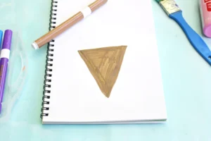 Open spiral notebook on a pastel blue desk with a brown triangular shape drawn in the center of the page and markers nearby.