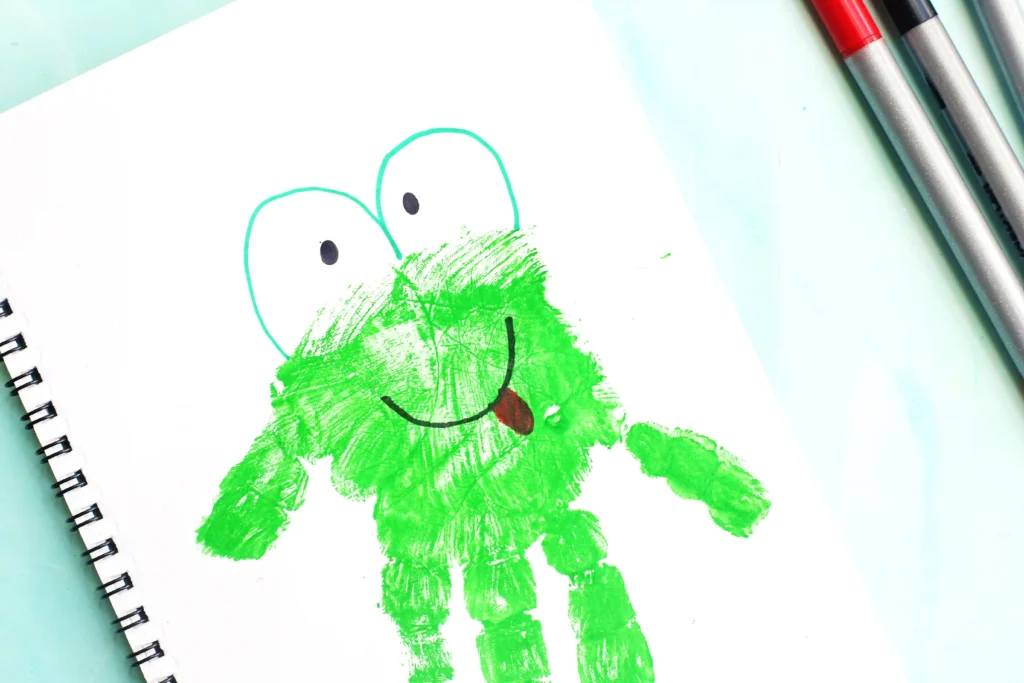 Letter F Frog Handprint Craft - Cultivating Brilliant Minds Childlike drawing of a green frog with big eyes and a red tongue on white paper, with markers nearby for a letter f craft