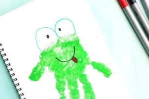 Childlike drawing of a green frog with big eyes and a red tongue on white paper, with markers nearby for a letter f craft