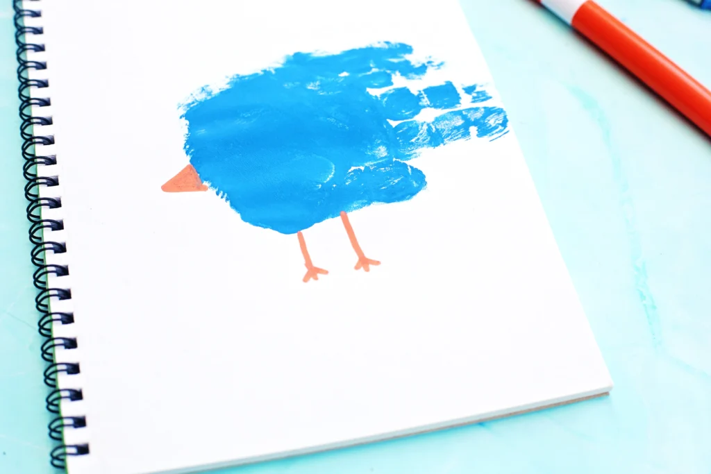White spiral notebook page featuring a blue bird silhouette with orange legs. The image has a bright blue paint mark on white paper and a small orange beak/feet detail nearby.